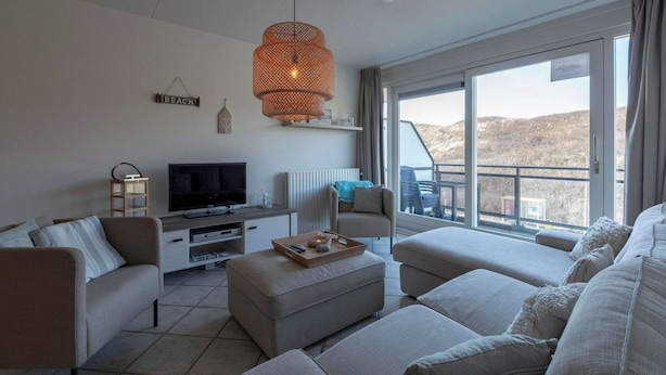 A modern living room with a flat-screen TV, a large window with a view of the hills, and a cozy seating area. - Wijde Blick 212 Apartment in Callanstoog (Callantsoog)