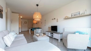 A living room with a sofa, a chair, a TV, and a dining area in the background. - Wijde Blick 212 Apartment in Callanstoog (Callantsoog)