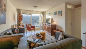 Un salon moderne comprenant un canapé, des fauteuils, une table basse et un téléviseur. Il y a un balcon avec vue sur la ville.