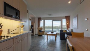 A modern kitchen with a built-in microwave, a sink, and a stove. There is a dining area with a table and chairs, and a sofa. The room has large windows with a view of the outdoors. - Wijde Blick 206 Apartment in Callanstoog (Callantsoog)