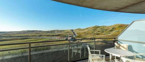 Ein Balkon mit Glastrennwand, weißen Gartenmöbeln und Blick auf eine Straße und Hügel.