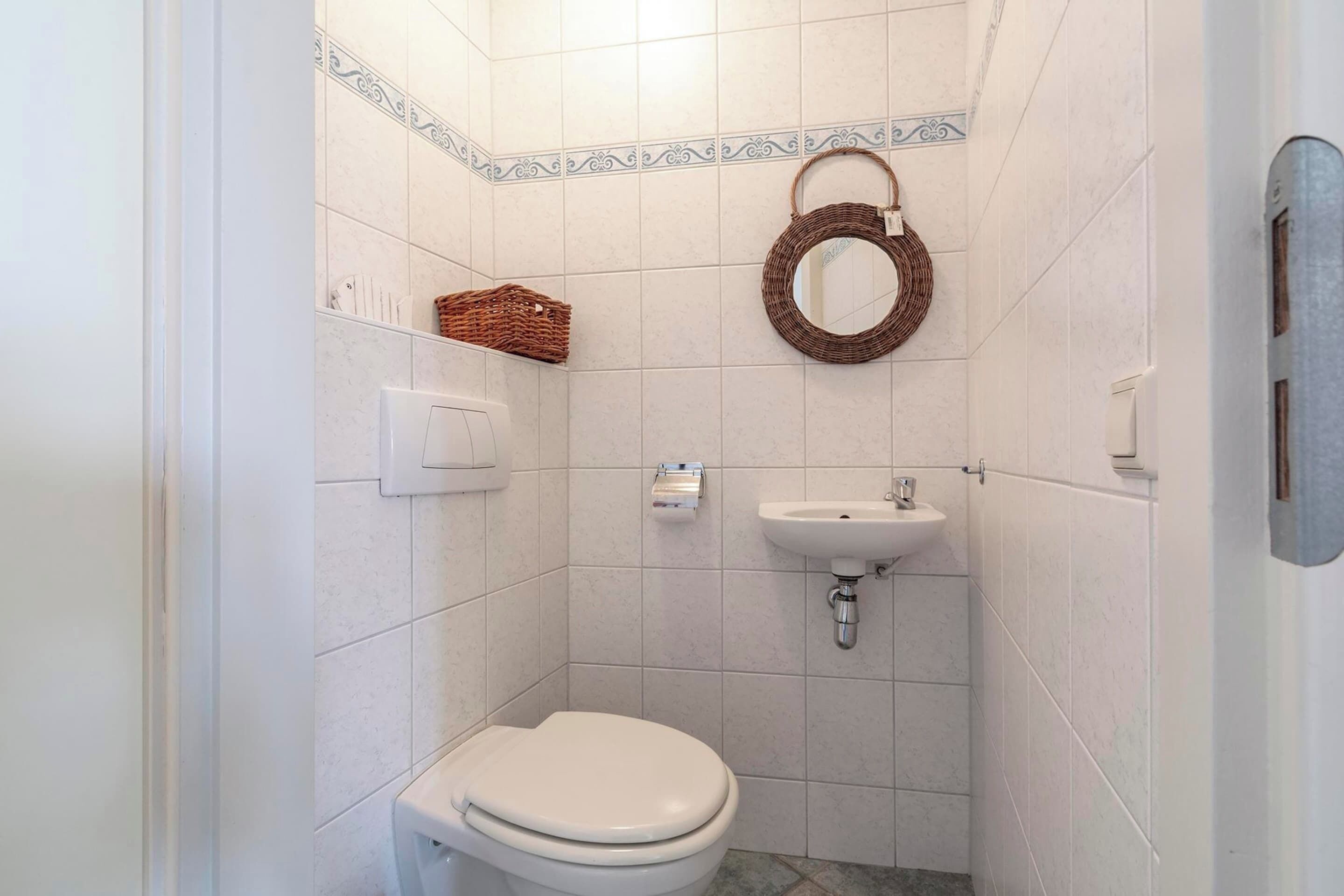 Un bagno piccolo e pulito, con piastrelle bianche, un water bianco, un lavandino e un cesto di vimini a parete.