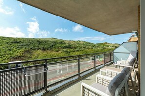 A balcony with a view of a green hillside, a glass railing, and outdoor seating with cushions. - Apartment in Callantsoog by the Beach (Callantsoog)