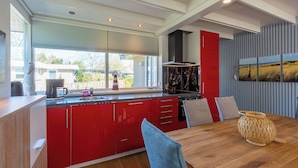 A modern kitchen with red cabinets, a wooden dining table, and a large window with blinds. - Modern Home Near Beach (Sint Maartensvlotbrug)