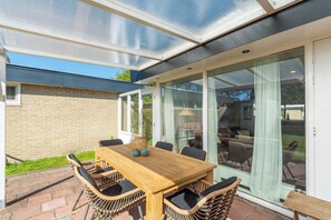 A patio with a wooden table and wicker chairs, a glass door leading to a garden, and a brick wall. - Modern Home Near Beach (Sint Maartensvlotbrug)