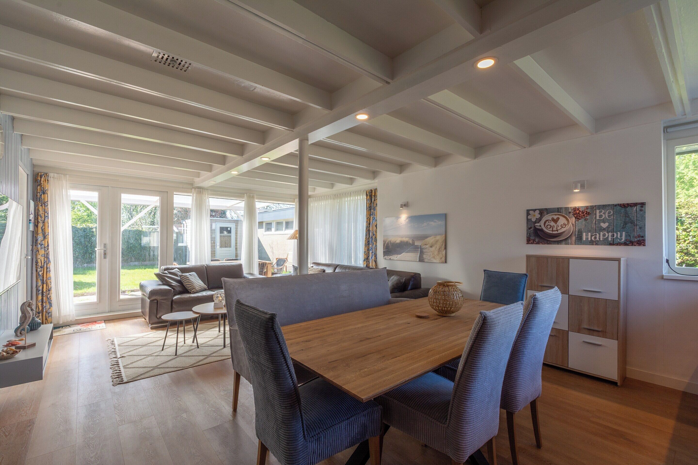 A spacious living area with a wooden dining table, blue upholstered chairs, a sofa, and a kitchen area with a cabinet and a decorative sign.