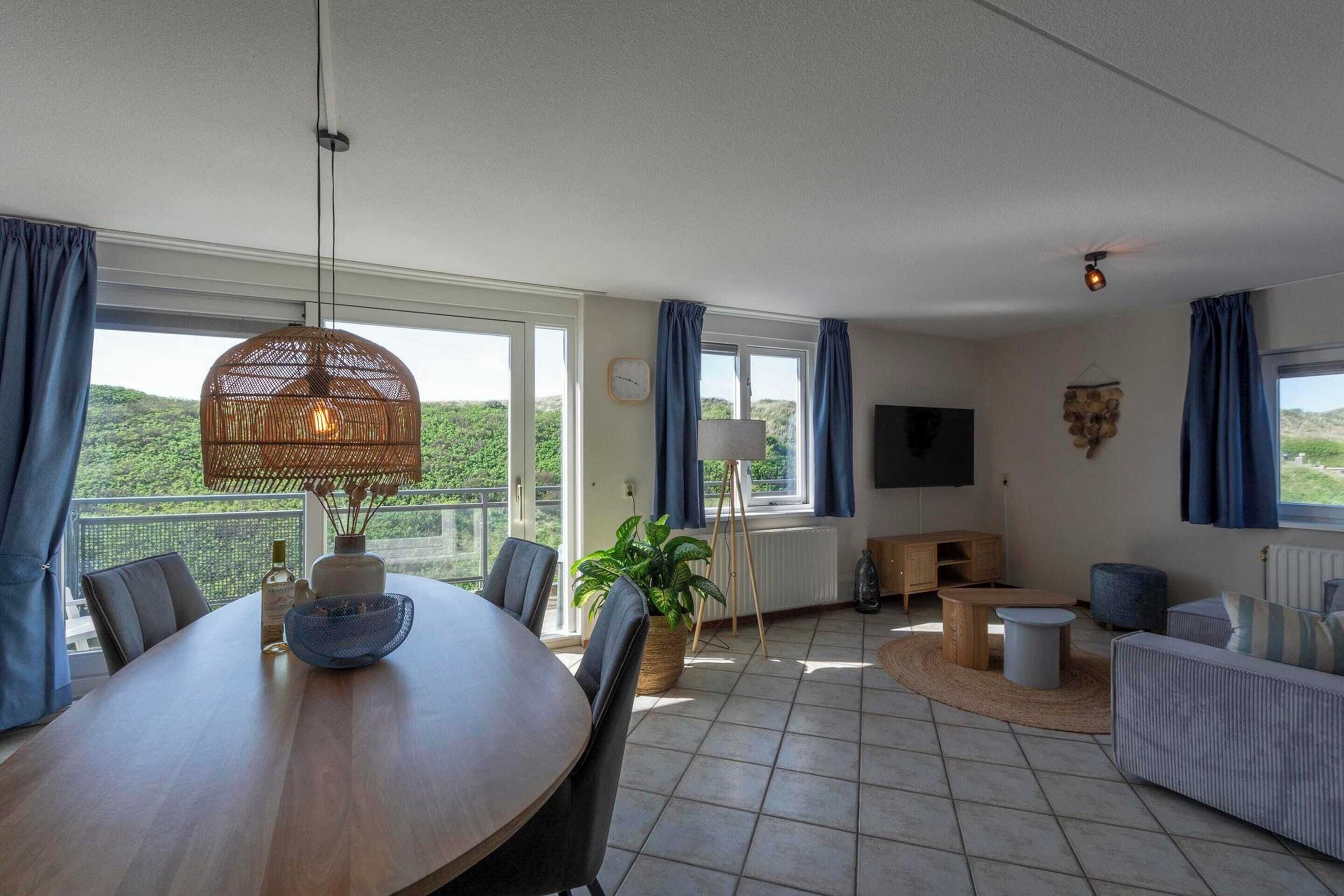 A spacious living area with a dining table, sofas, and a TV. Large windows offer a view of the outdoors.