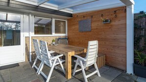 A covered patio with a wooden table and white chairs, a wooden wall with a decorative panel, and a window with blinds. - Wildrijk 83 Holiday Home Near Beach (Sint Maartensvlotbrug)
