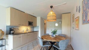 A modern kitchen with light wood cabinets, a central island, and a dining area with a round wooden table and grey chairs. - Wijde Blick 310 Apartment Near Callanstoog Beach (Callantsoog)
