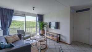 A modern living room with a sofa, chairs, a TV, and a large window with curtains. - Wijde Blick 310 Apartment Near Callanstoog Beach (Callantsoog)