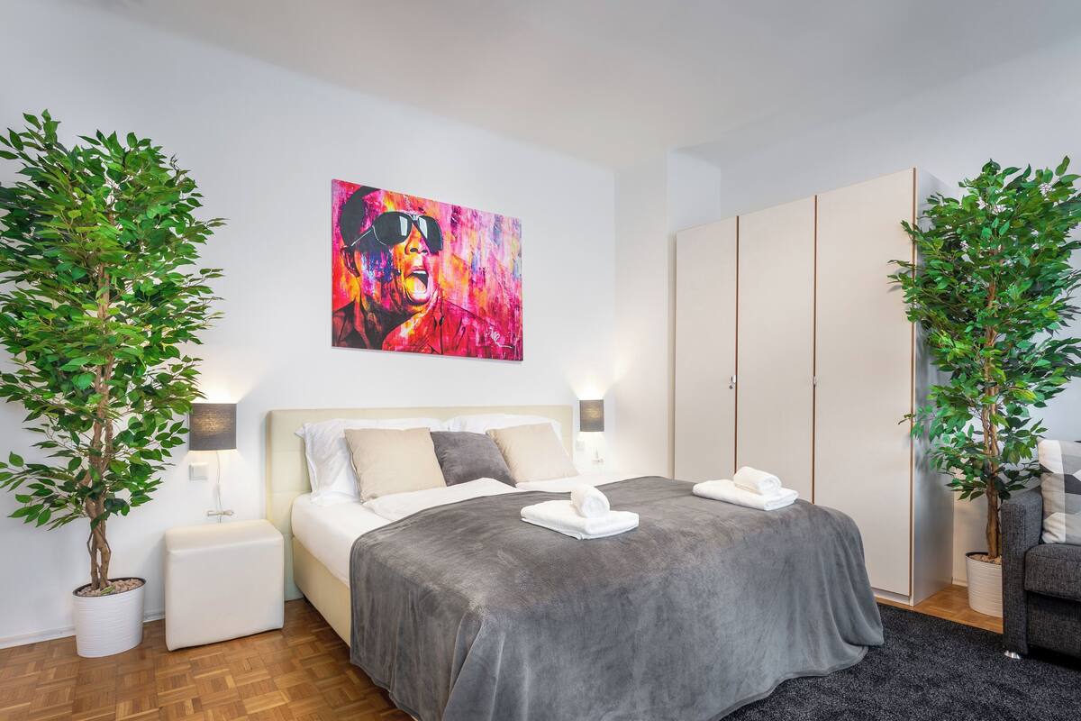 A modern bedroom with a bed, bedside tables, a painting, and two potted plants.
