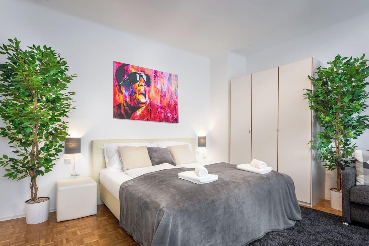 Une chambre à coucher moderne avec un lit, des tables de chevet, un tableau et deux plantes en pot.