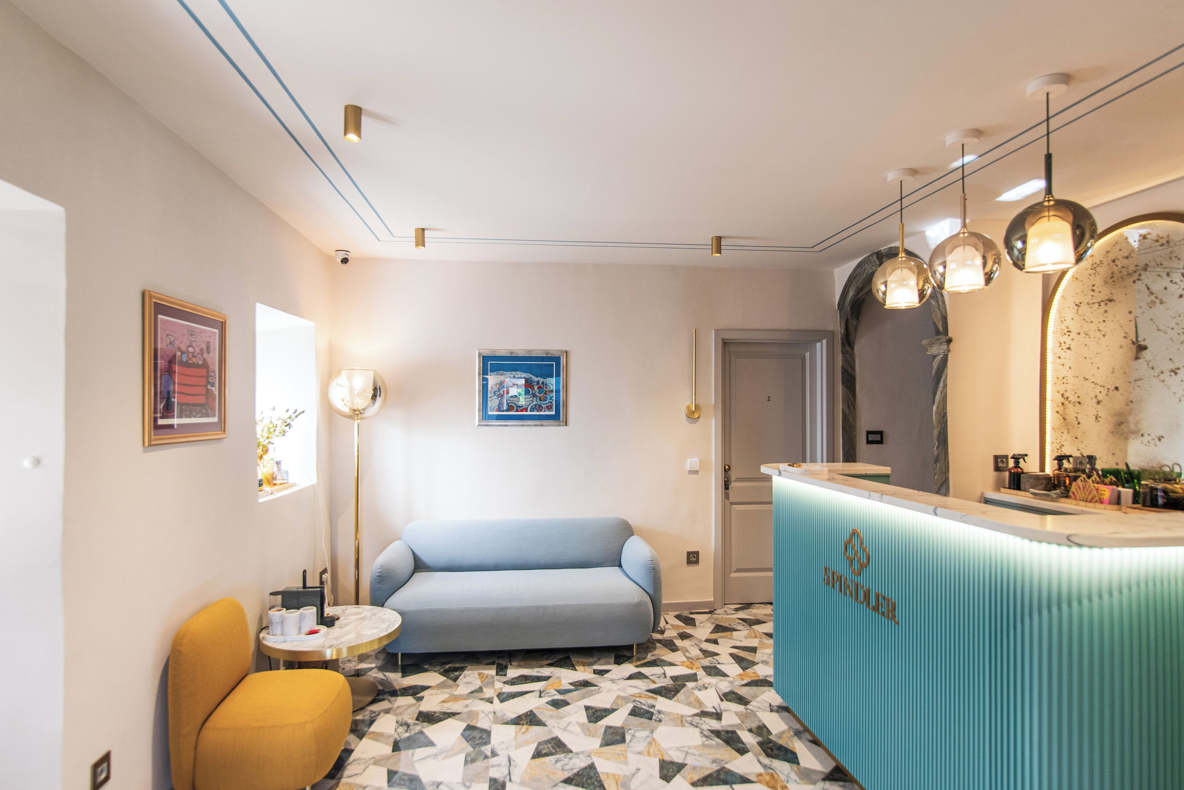 A modern hotel room with a blue sofa, a yellow armchair, a small round table, and a reception desk with a blue counter.