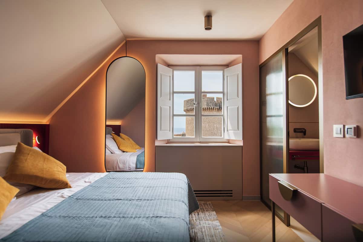 Un dormitorio con cama, ventana con vista a un edificio, espejo y mesita de noche.
