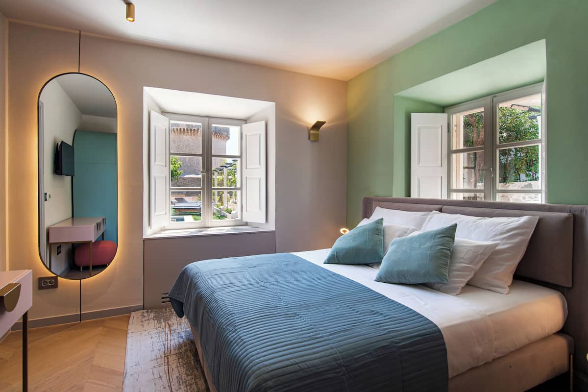 Un dormitorio con una cama, un espejo, una ventana y una lámpara en la pared.