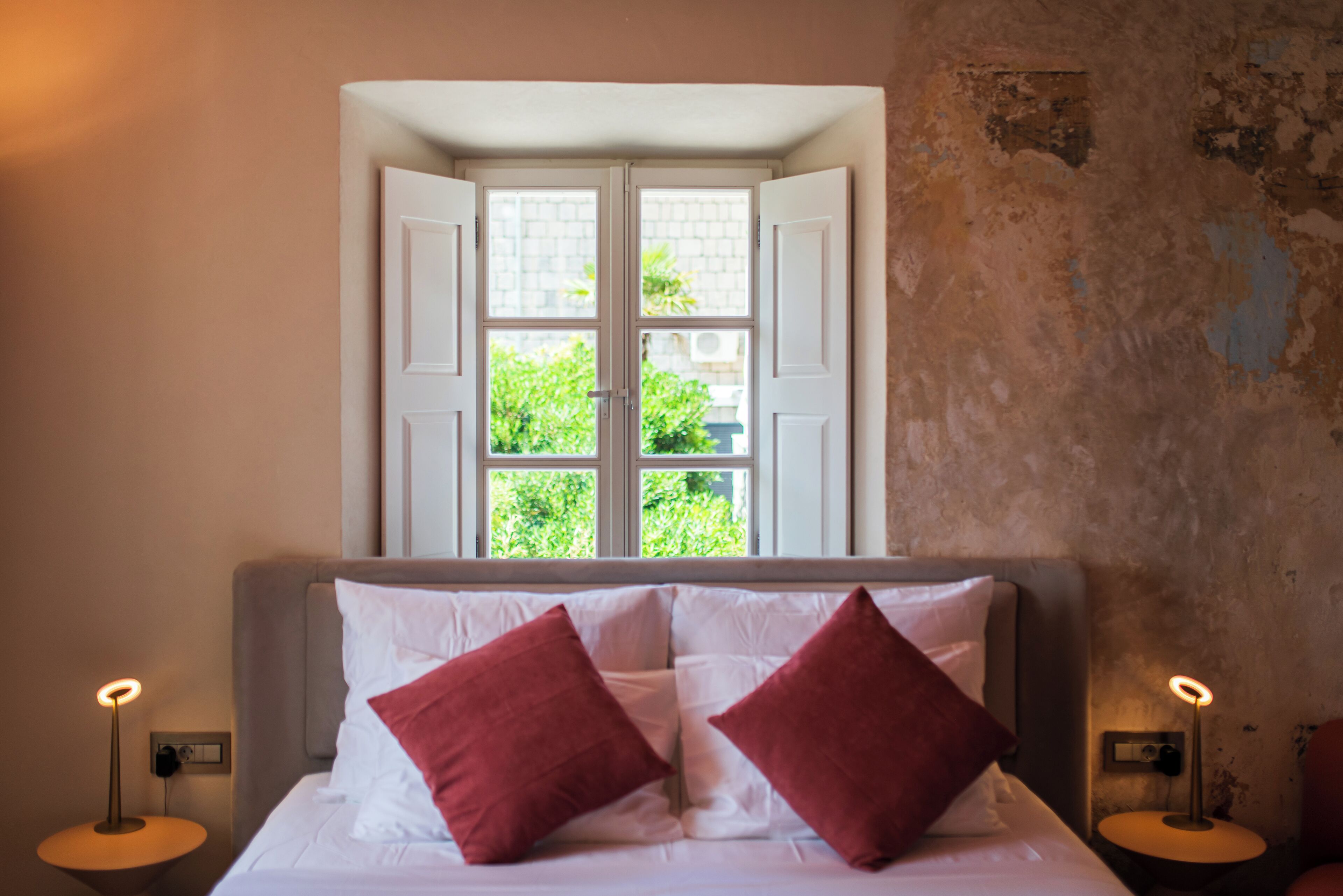 Ein Schlafzimmer mit einem Bett, zwei roten Kissen, einem Fenster mit geöffneten Fensterläden und Blick auf Grün.