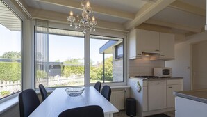 Une cuisine moderne avec des meubles blancs, une table à manger avec des chaises, un lustre et une vue sur un jardin grâce à de grandes fenêtres.