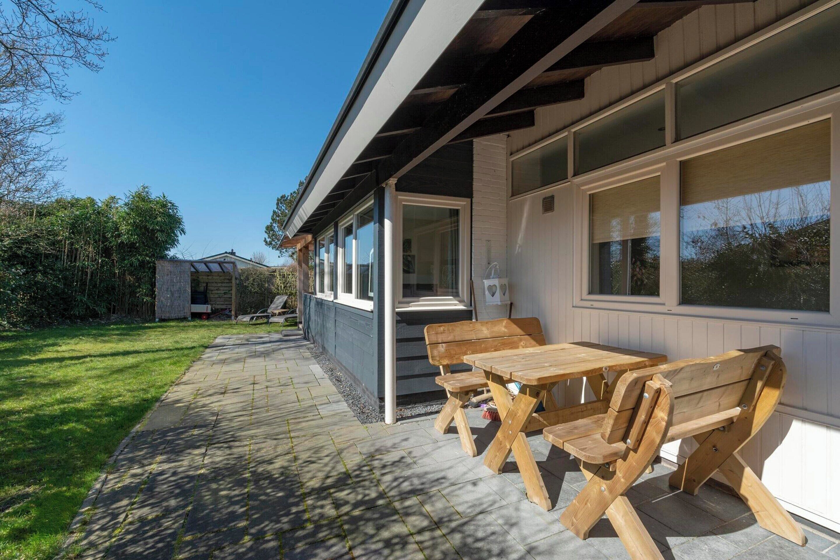 A covered outdoor seating area with wooden benches and tables, a stone pathway, and a view of a house with large windows and a covered porch.