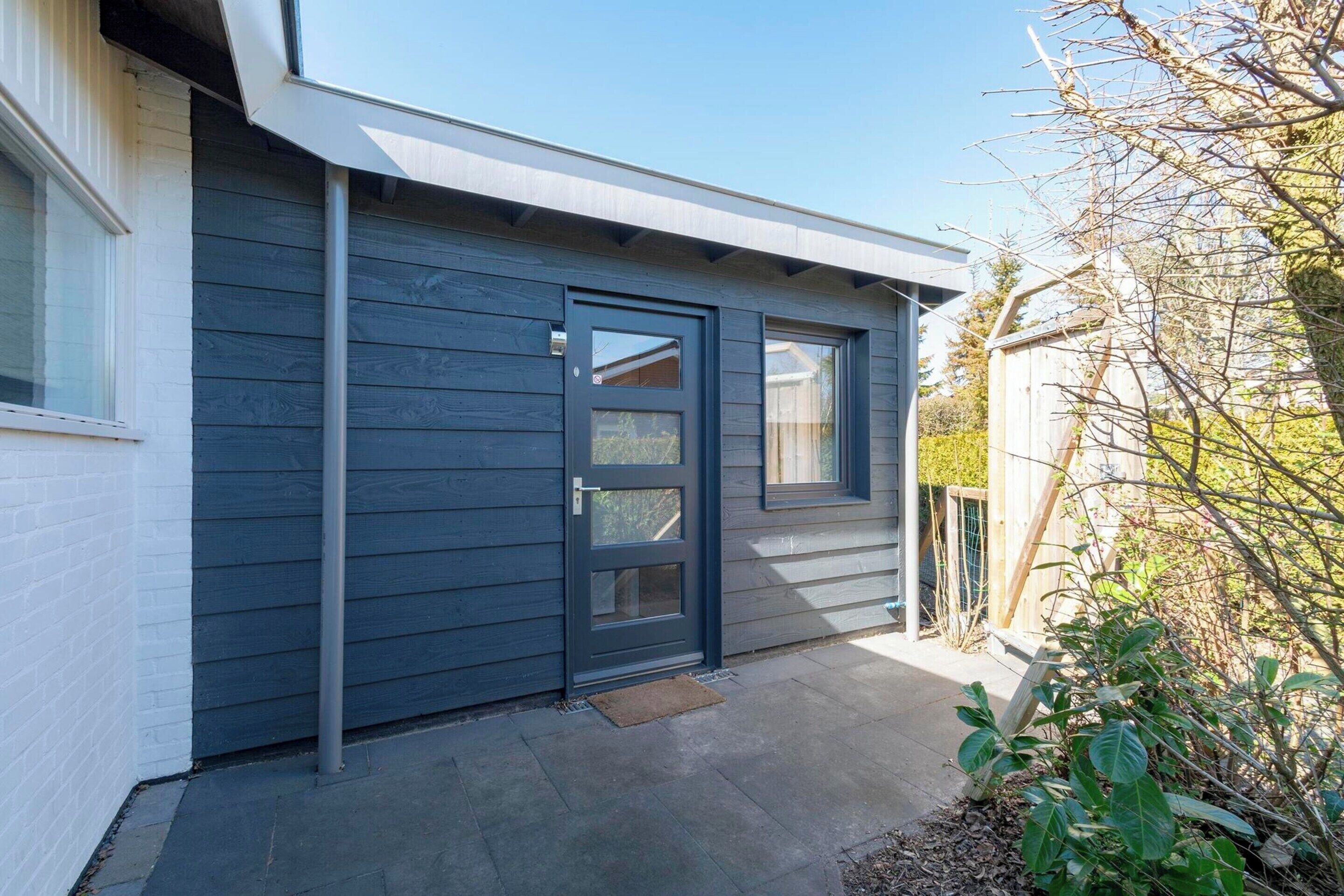 Une maison moderne avec une façade de couleur gris foncé, une porte d’entrée dotée d’un panneau de verre et un petit abri en bois à l’arrière.