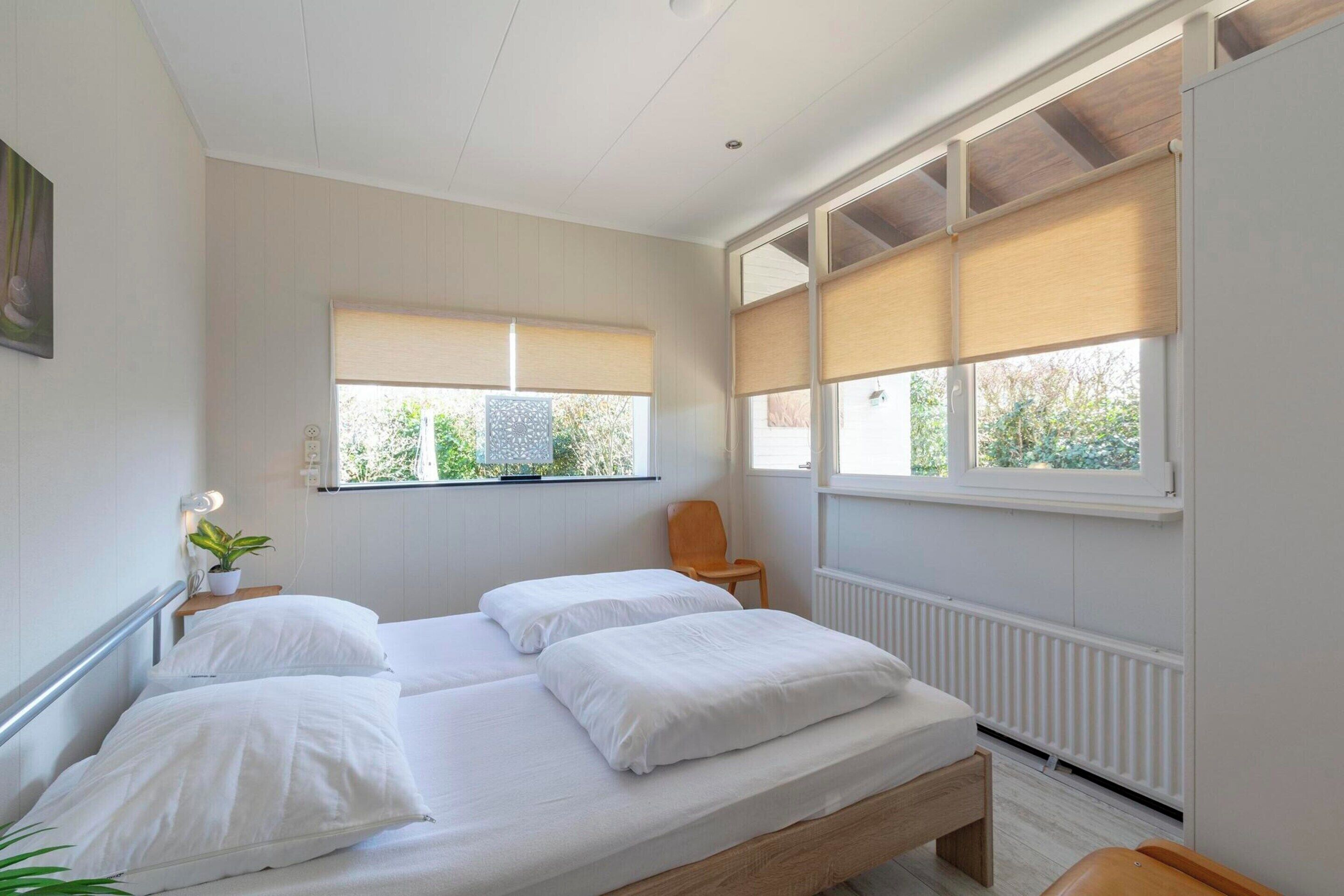 A bedroom with two beds, a chair, a potted plant, and large windows with blinds.