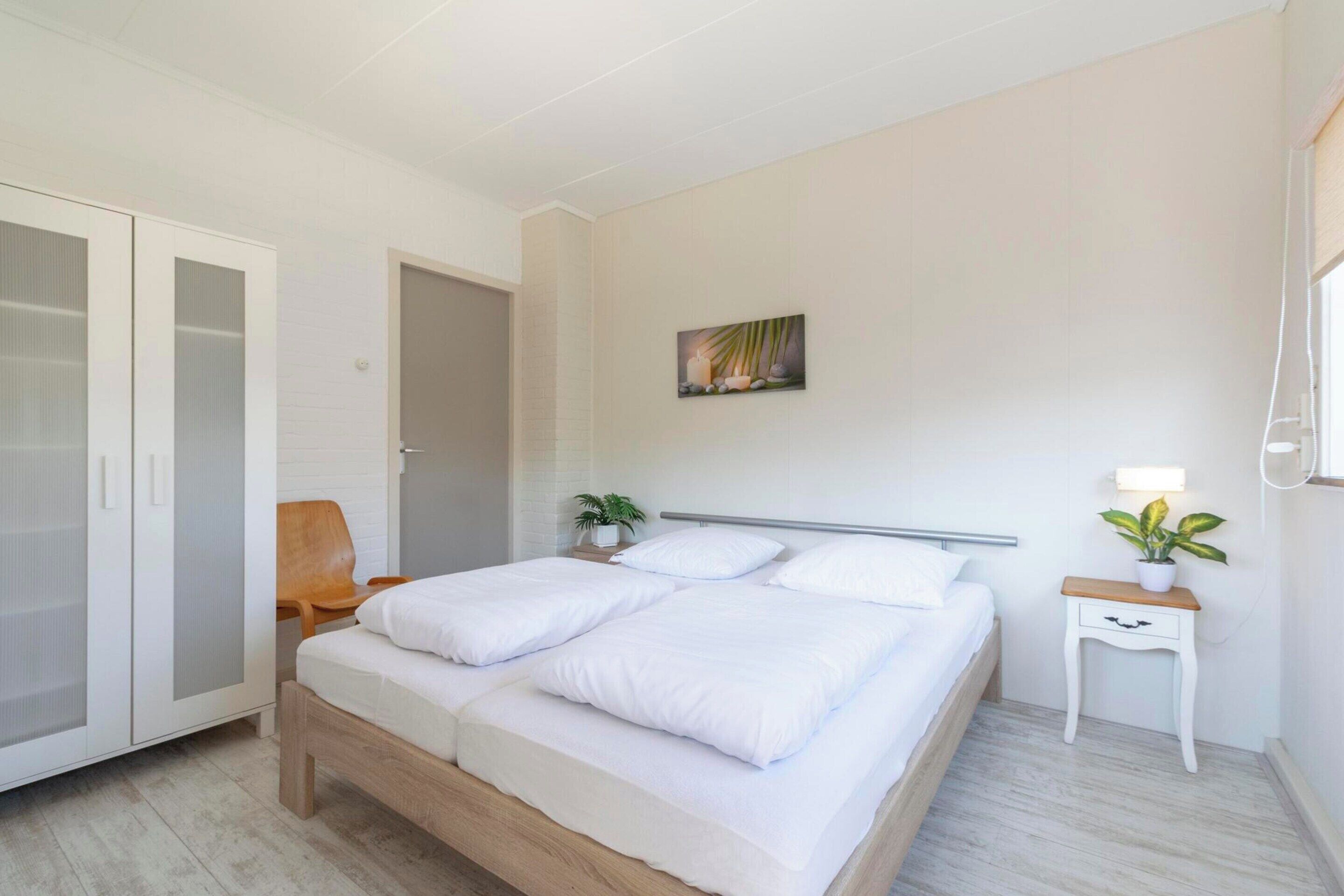 A bedroom with a bed, a wardrobe, a chair, a bedside table with a lamp, and a wall-mounted artwork.