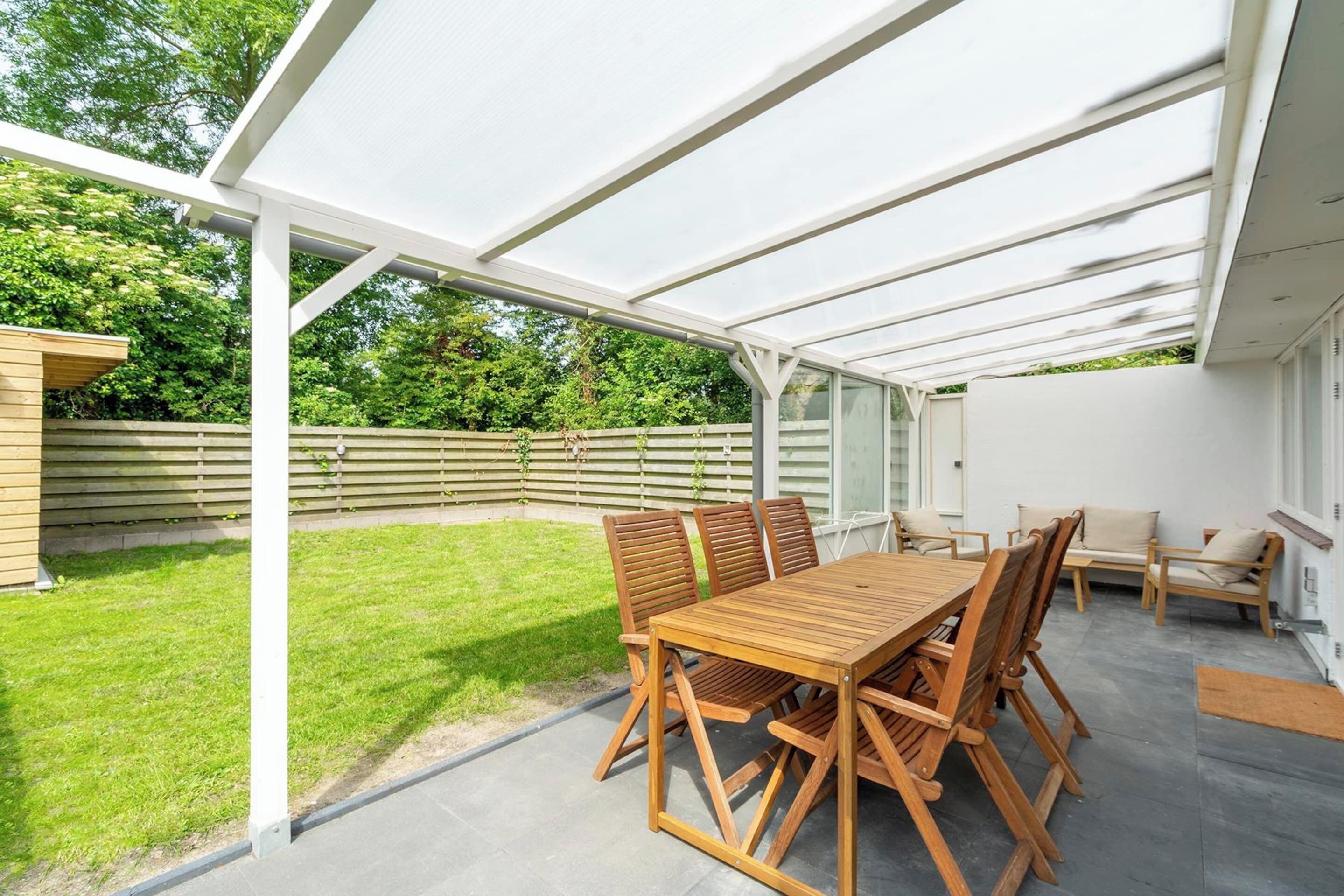 A covered patio area with a wooden table and chairs, a glass enclosure, and a view of a green garden.