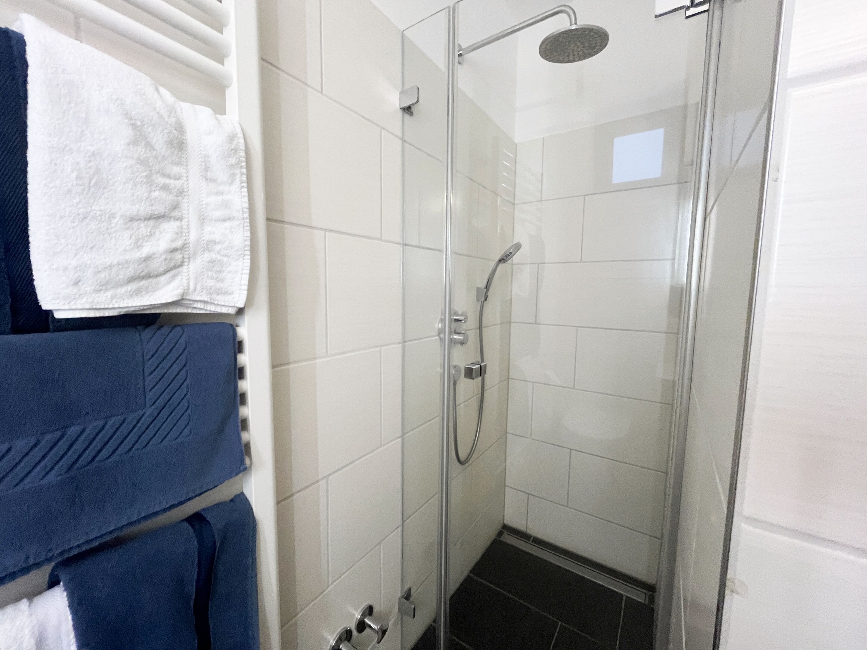 Ducha con paredes de azulejos blancos, cabezal de ducha de mano y toallero con toallas blancas y azules.