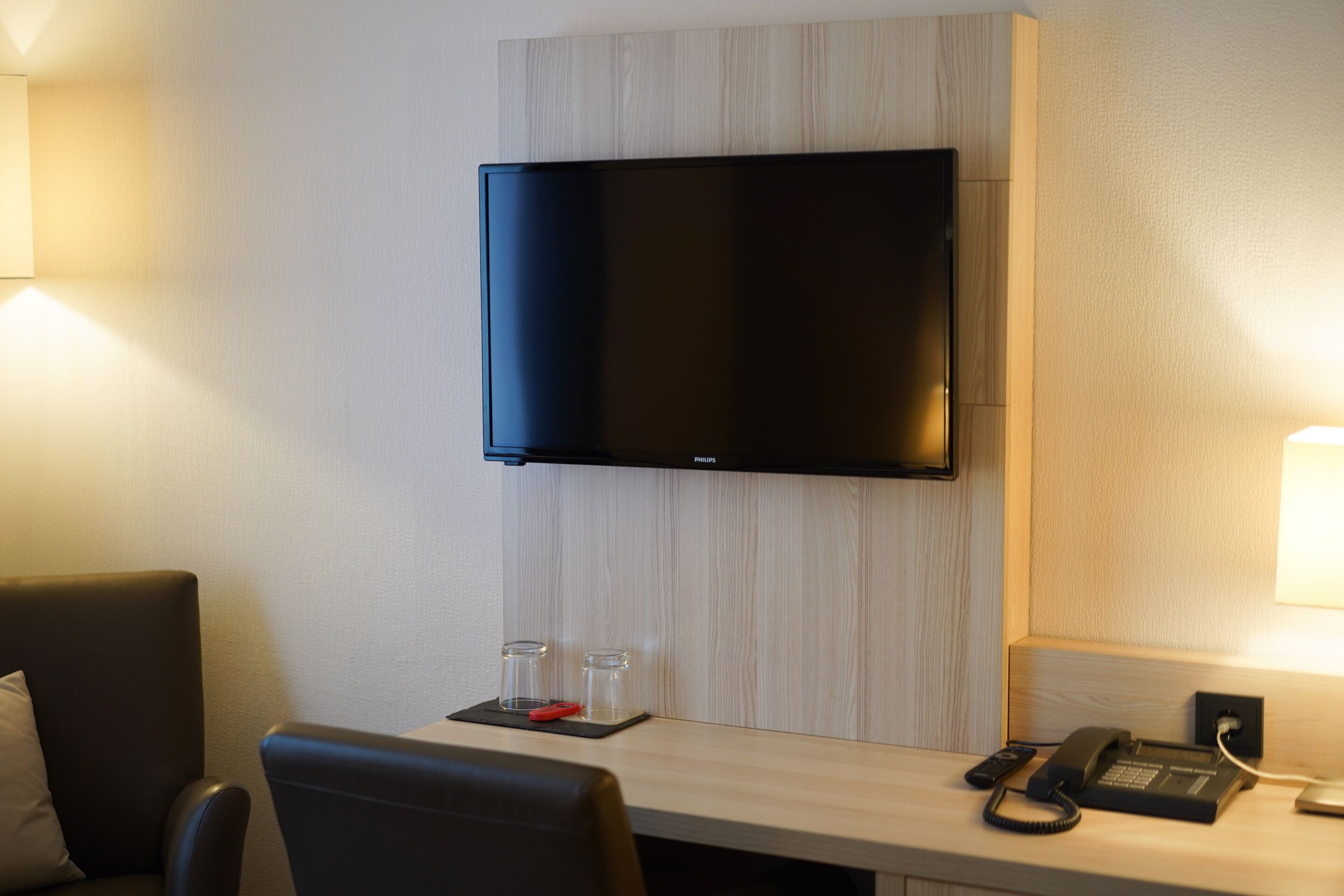 Habitación de hotel con televisión de pantalla plana montada en la pared, un escritorio con teléfono y control remoto, y dos pequeños frascos de vidrio sobre un posavasos.