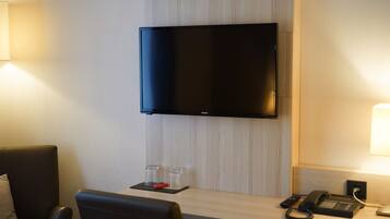 Habitación de hotel con televisión de pantalla plana montada en la pared, un escritorio con teléfono y control remoto, y dos pequeños frascos de vidrio sobre un posavasos.