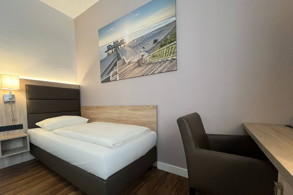 A hotel room with a bed, a desk, and a painting of a pier on the wall.
