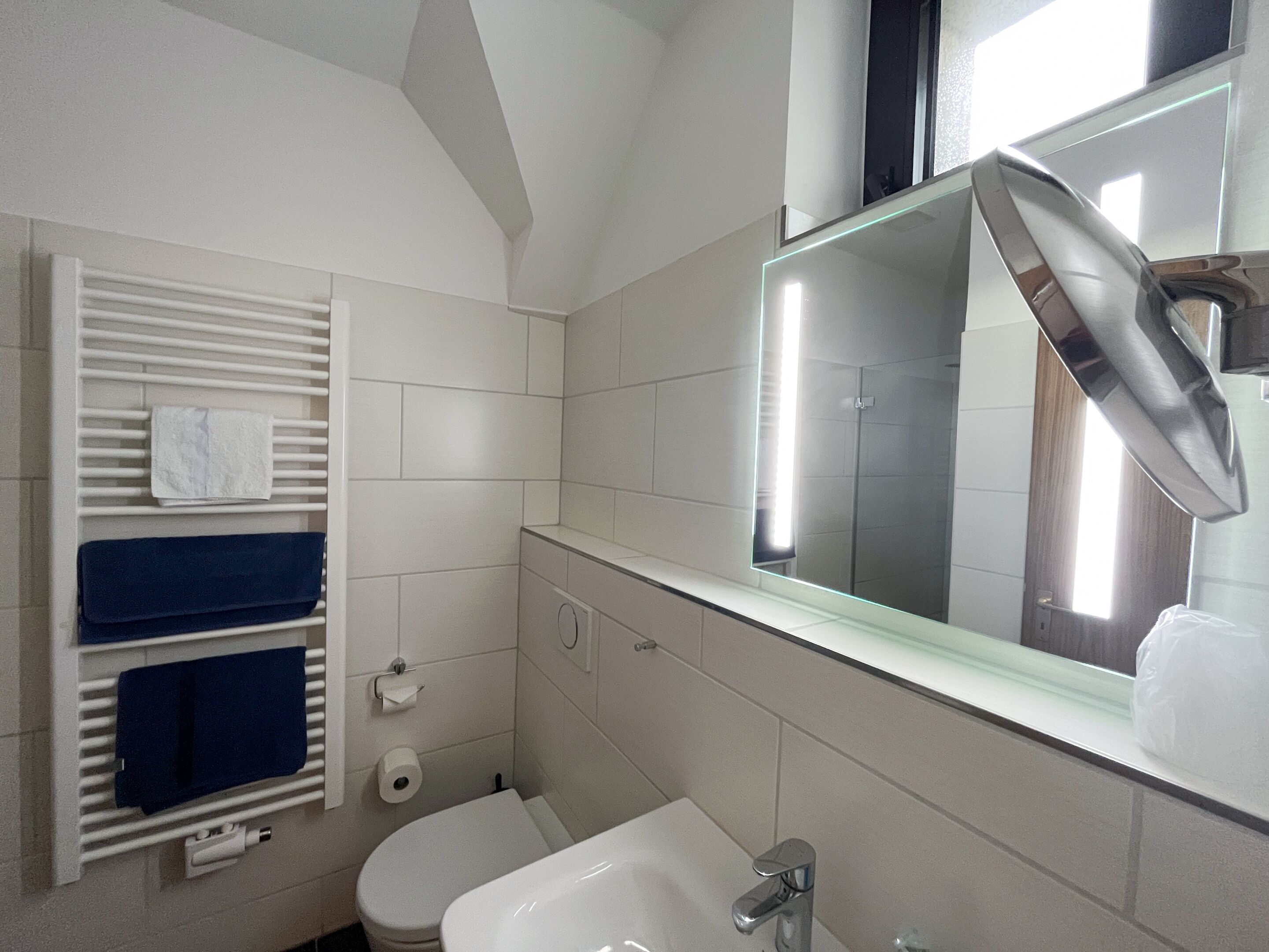 Un baño con inodoro, lavamanos y espejo. Hay un perchero con toallas y un dispensador de papel higiénico.