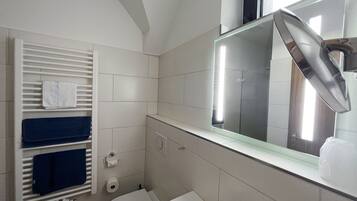 Un baño con inodoro, lavamanos y espejo. Hay un perchero con toallas y un dispensador de papel higiénico.