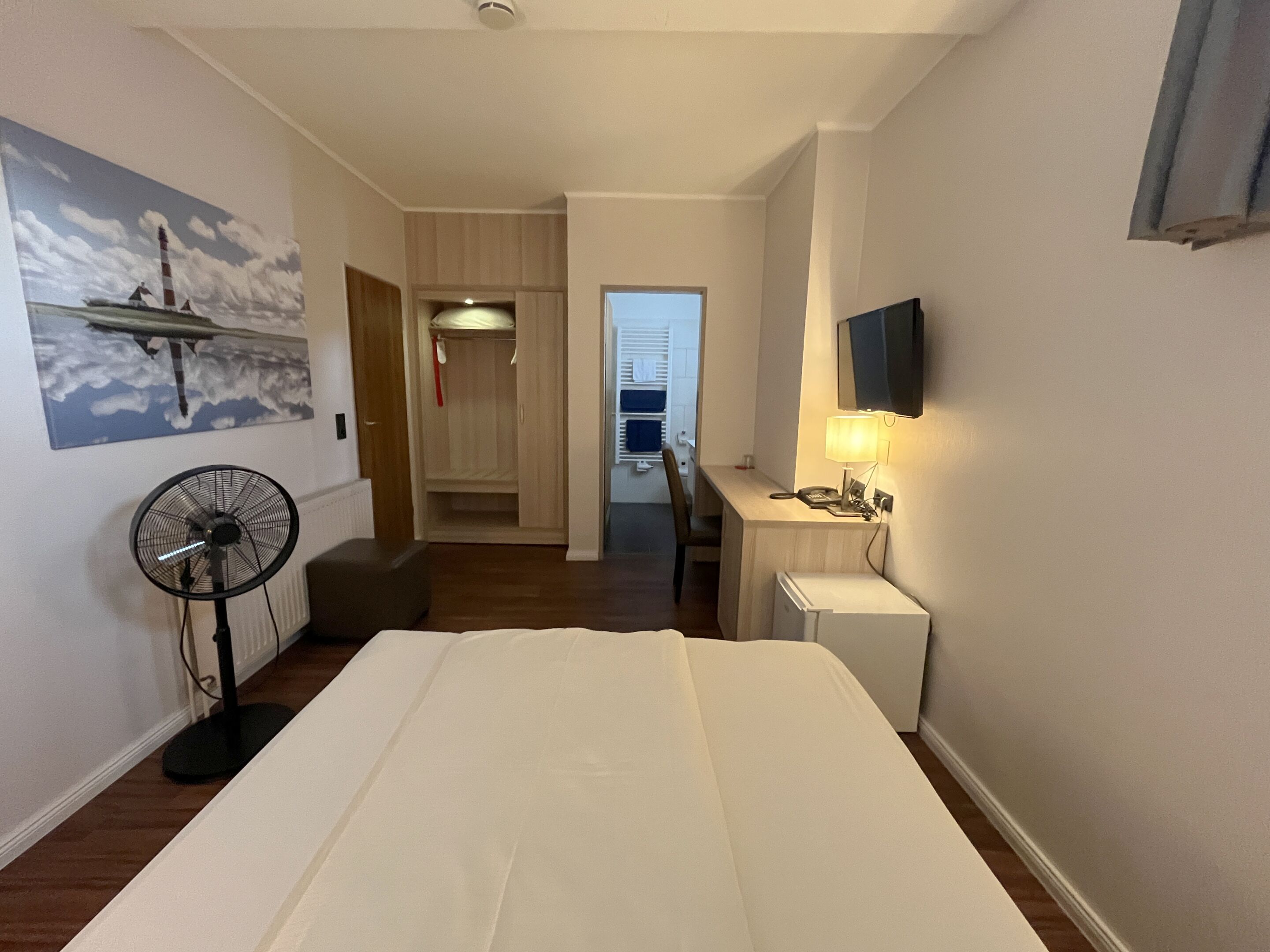 Habitación de hotel con cama, escritorio con lámpara, televisión, ventilador y armario.
