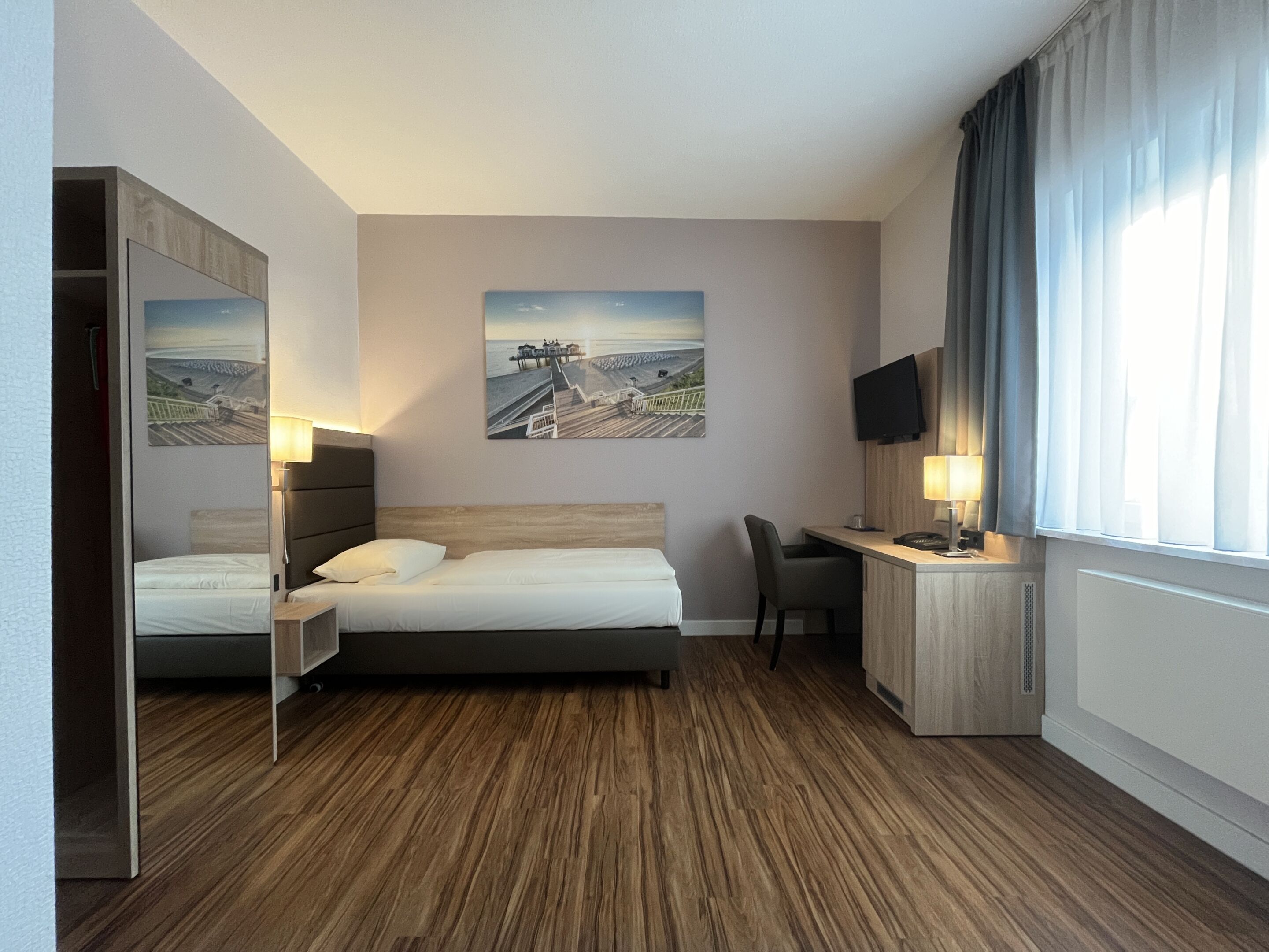 A hotel room with a bed, a desk, a chair, a TV, and a painting on the wall.