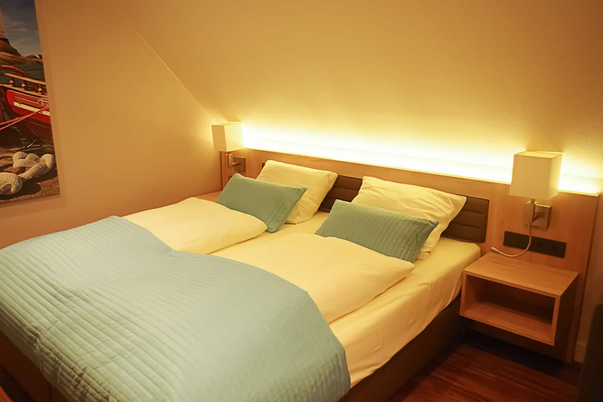 A neatly made bed with a quilted green blanket and two pillows, a bedside table with a lamp, and a wall-mounted shelf.