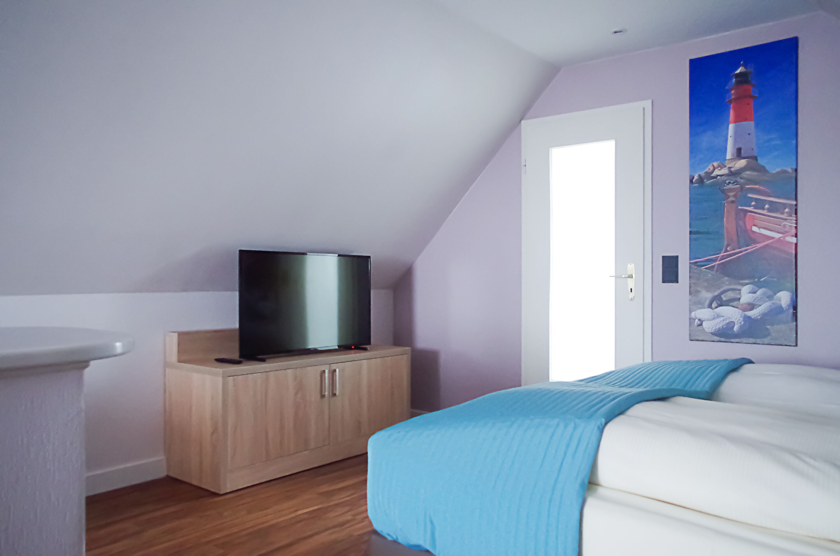 A bedroom with a bed, a TV on a wooden cabinet, and a painting of a lighthouse on the wall.