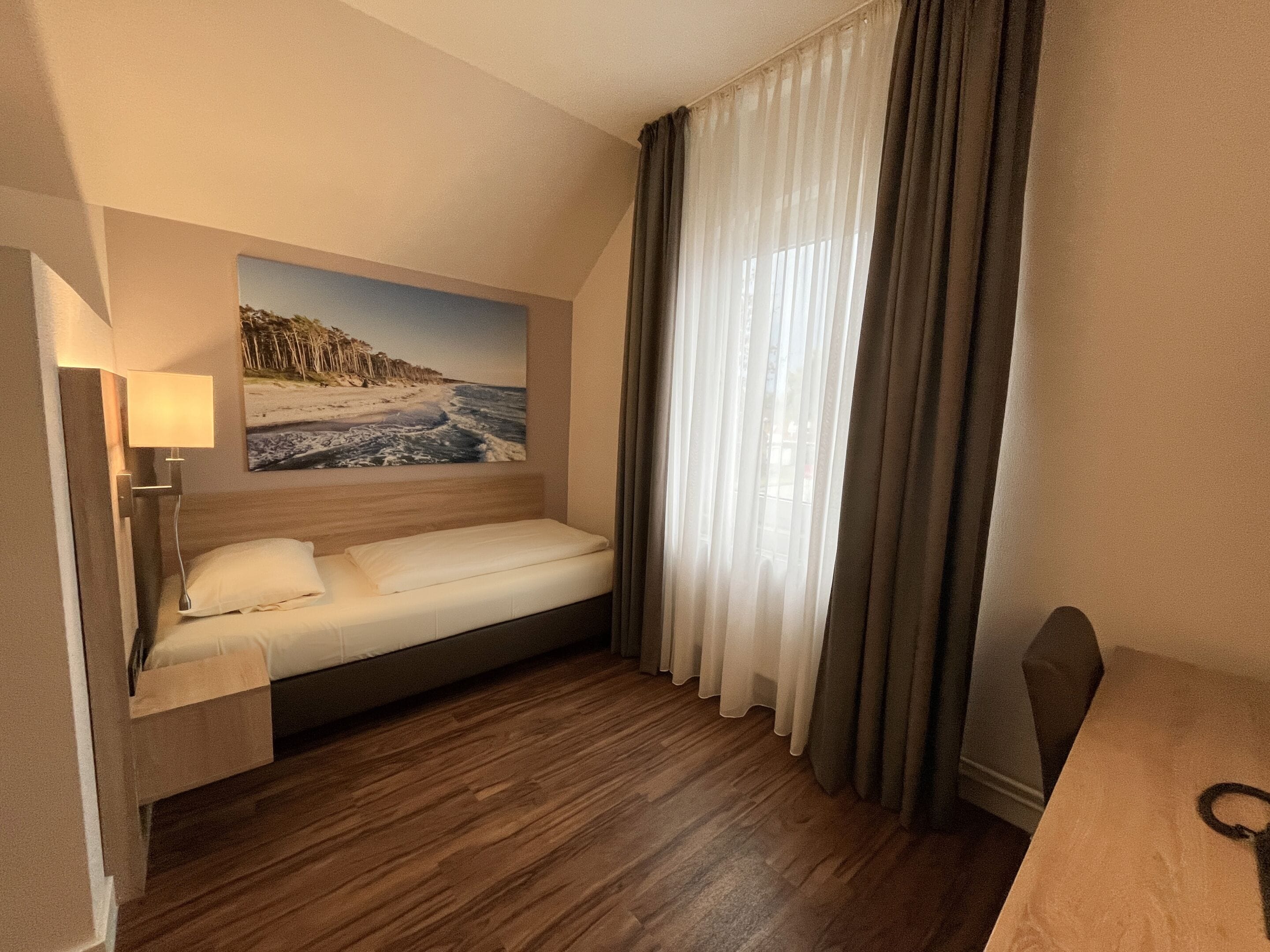 A hotel room with a bed, a nightstand, a lamp, a painting of a beach scene, and a window with curtains.