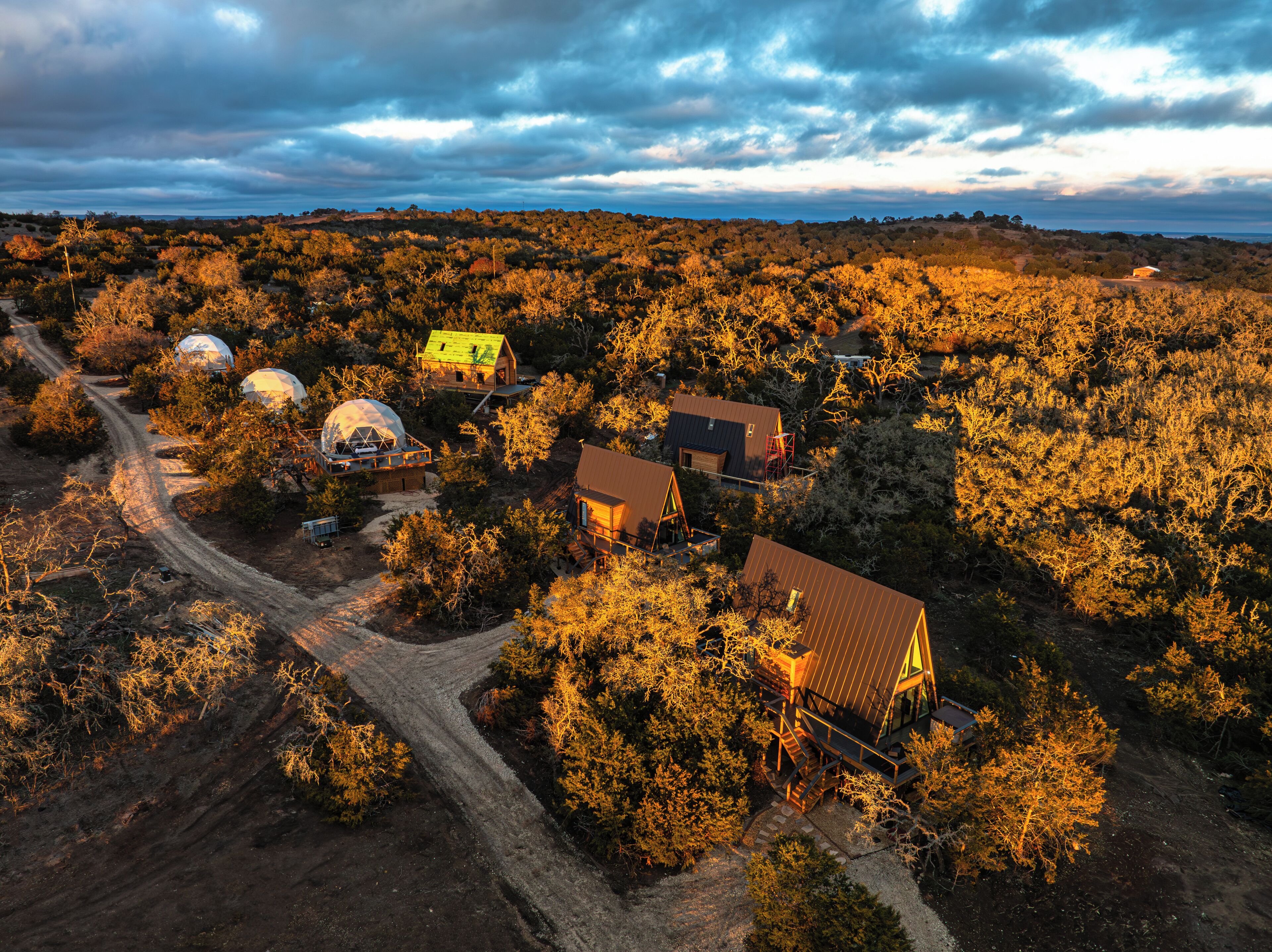 An aerial view of a rural area with several dome-shaped structures and a few traditional buildings, surrounded by dense vegetation and a dirt road.