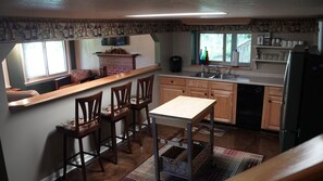 A kitchen with wooden cabinets, a dining table, and chairs. There is a window with a view of greenery outside.