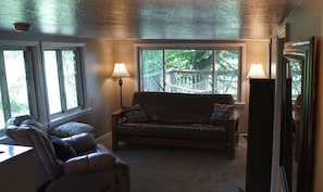 A cozy living room with a leather sofa, two armchairs, and a wooden coffee table. There are two floor lamps and a large window with a view of a wooden deck and trees.