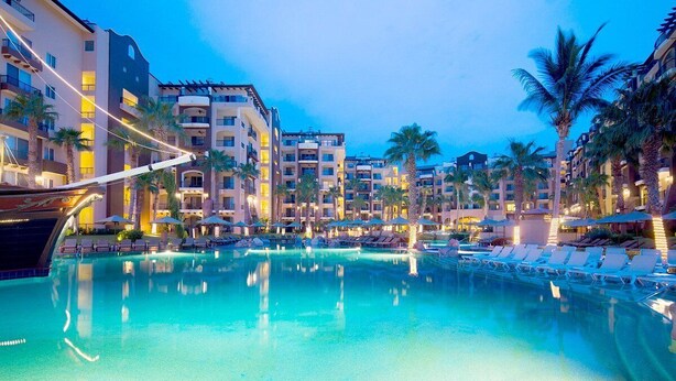 Ein Hotelpoolbereich mit mehreren Liegestühlen und Sonnenschirmen, umgeben von Palmen und mehrstöckigen Gebäuden.