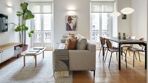 Uma sala de estar moderna com um sofá, uma mesa de jantar com cadeiras, uma televisão e uma janela grande com vista para edifícios.