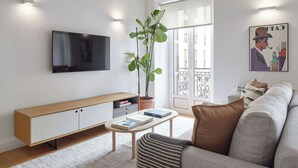 Uma sala de estar moderna com uma televisão de tela plana, um console de madeira, uma mesa de centro redonda, uma almofada listrada, um sofá cinza e uma planta grande em vaso.