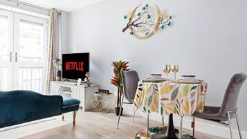 Moderni olohuone, jossa on litteänäyttöinen televisio, jonka näytöllä näkyy teksti »NETFLIX», ruokapöytä, jonka päällä on lehtikuviollinen pöytäliina, sekä koristeellinen seinätaulu pöydän yläpuolella.