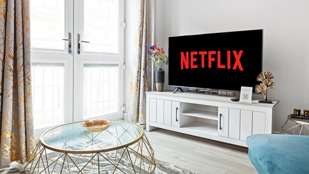 Un salon avec un téléviseur affichant le logo « NETFLIX », une table basse en verre, un meuble de rangement blanc et un canapé bleu.