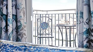 Un balcon avec une chaise en métal et une table à motifs de mosaïque, des rideaux à fleurs et une vue sur le paysage urbain.