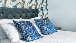 Un lit recouvert d’une couvre-lit blanche à texture, agrémenté de deux coussins bleus à motifs, doté d’une tête de lit grise à motifs en relief et placé devant un mur orné d’un motif évoquant le marbre.