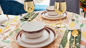 Une table à manger dressée avec de l’assiette blanche, des couverts dorés et deux verres à vin dorés.