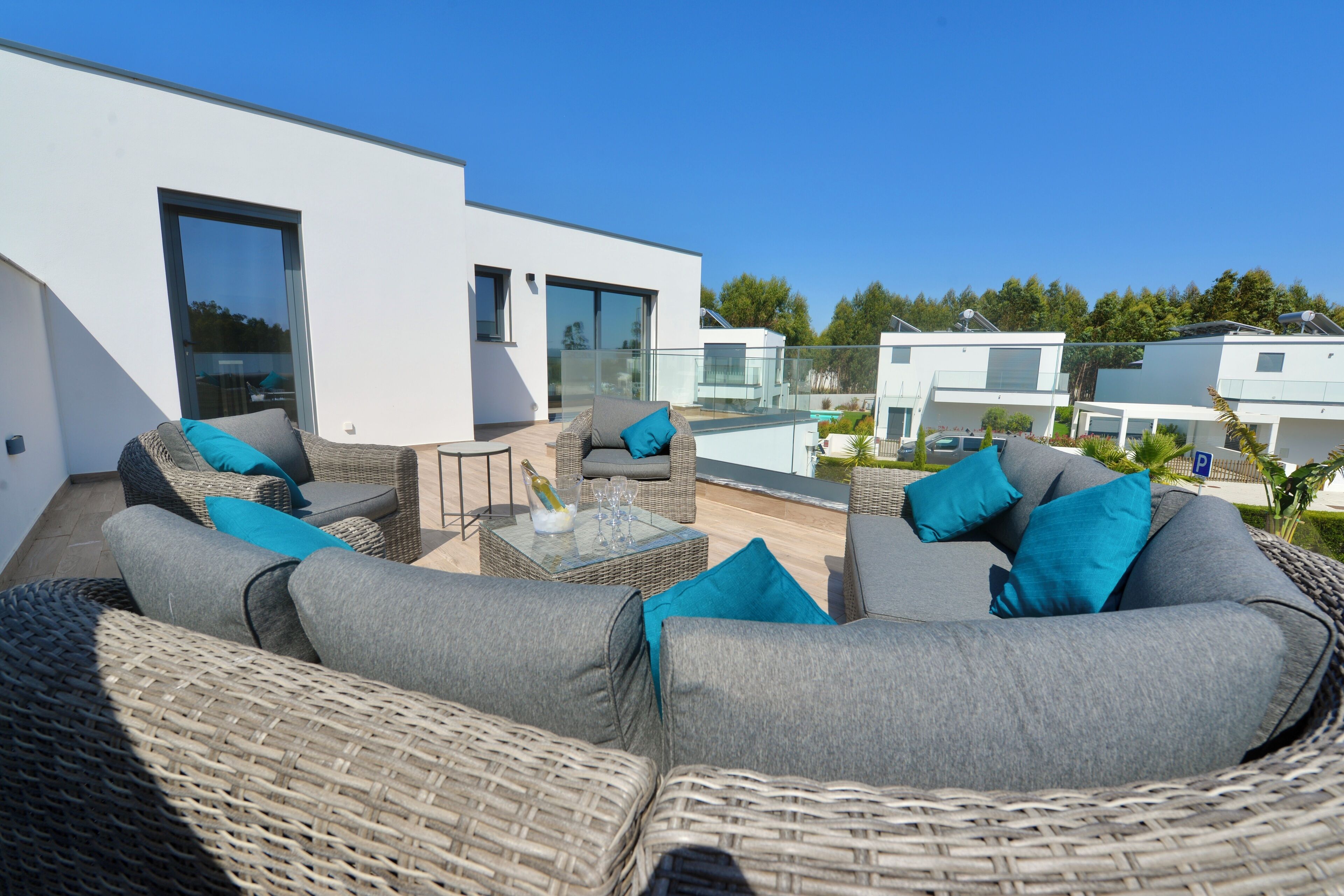 Een moderne buiten zithoek met rieten meubels en kussens, een glazen tafel met een fles en glazen, en een helder uitzicht op een wit gebouw en groen in de achtergrond.