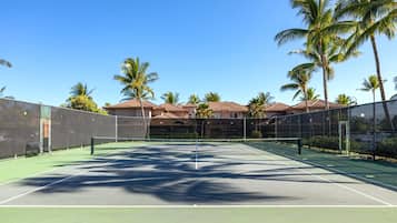 A tennis court with a net, surrounded by a fence, palm trees, and a building with a red roof in the background.